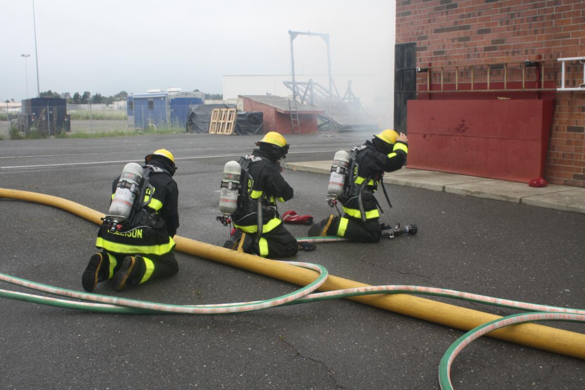 Fire fighter recruits in full turnout gear work with hoses out side of the training building