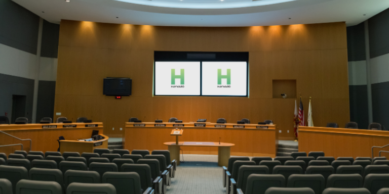 Photo of Hayward Council Chambers. A large auditorium-type room with audience seat and a Dais for the Council. 