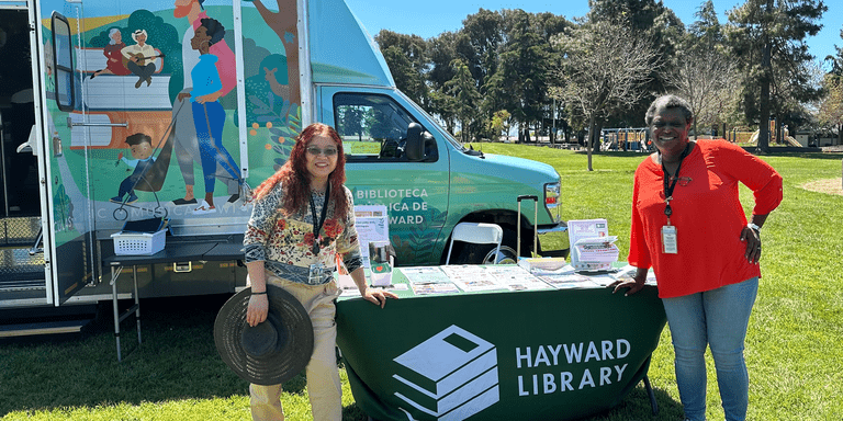 Two members of library staff standing near a library table in front of Curbie, the mobile library branch at an event in a park