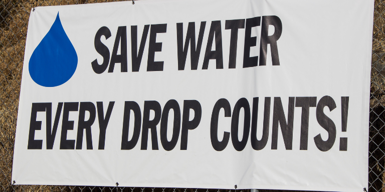 A white fence sign black text next to a blue water drop reads: Save Water Every Drop Counts!