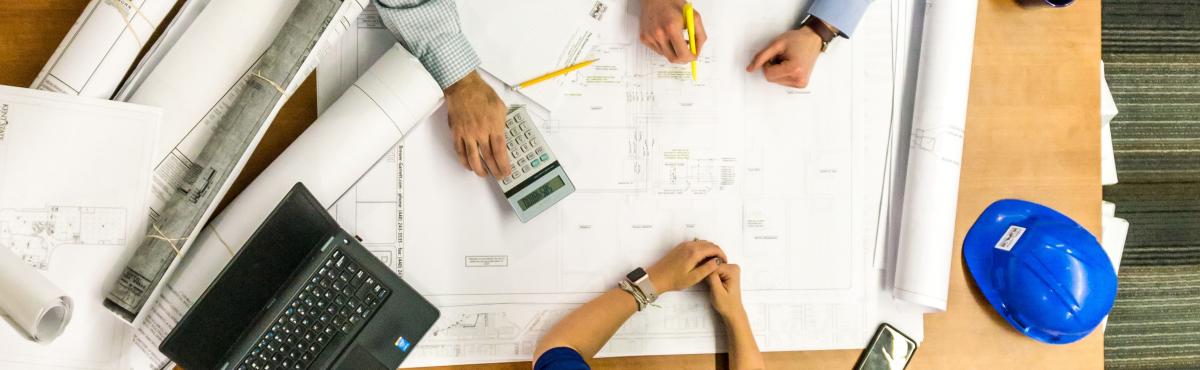 two people working at an architect's table. there are building plans and a blue hard hat on top.