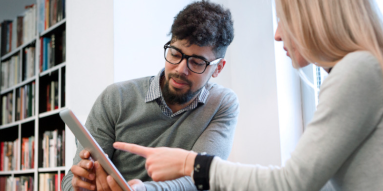 A tutor helps a learner use a tablet device with books in the background