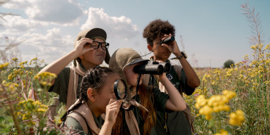 A group of children in an open field using telescopes, binoculars, and a magnifying glass