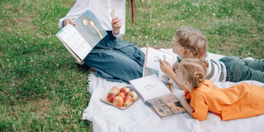 Children reading books on a picnic blanket in an open field. A caregiver is also present reading a book to them.