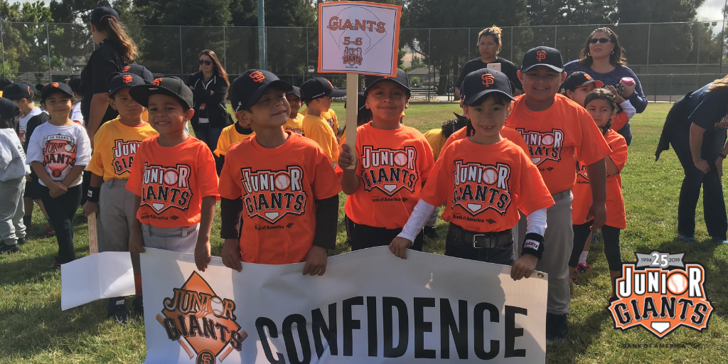 A group of smiling children wearing black and orange San Francisco Giants baseball hats and Junior Giants tee shirts holding a white sign that says confidence