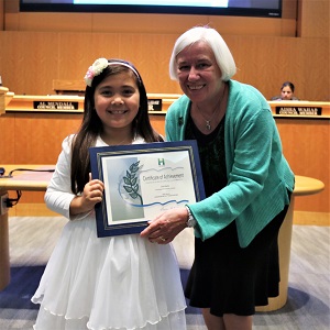 Little girl in a white dress with a white head band standing with Mayor Halliday