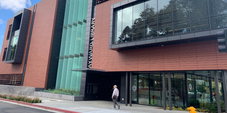 Image of a person walking in front of the Hayward Library, a large, dark red building with glass doors and windows throughout.