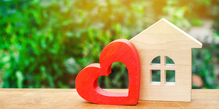 Photo of a small wooden house figurine next to a red wooden heart figurine on a wooden table. 