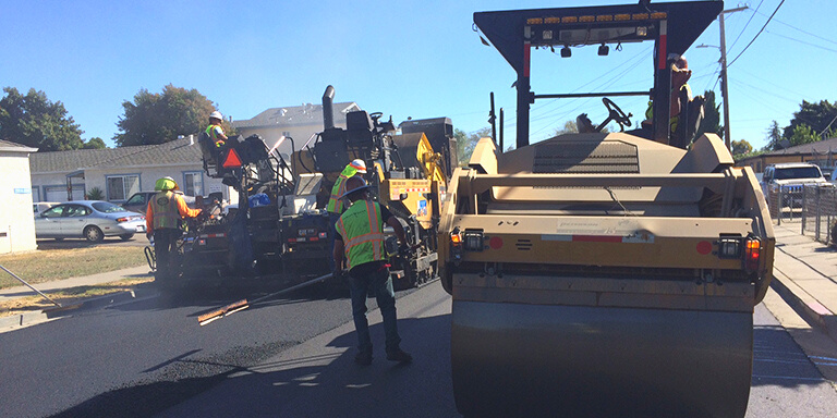 People in safety vests working on repaving the left side of a road with large pavement equipment.