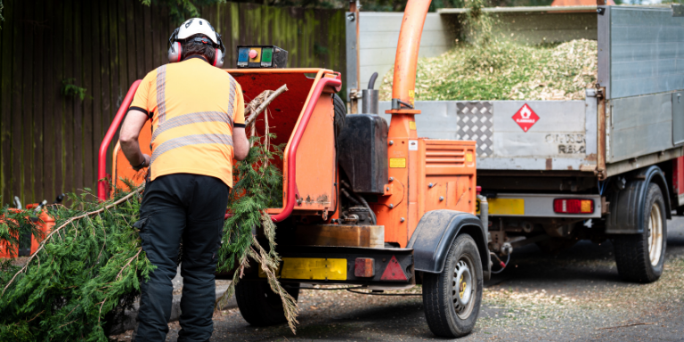 A person with proper protective gear feeding a tree branch into an orange woord chipper